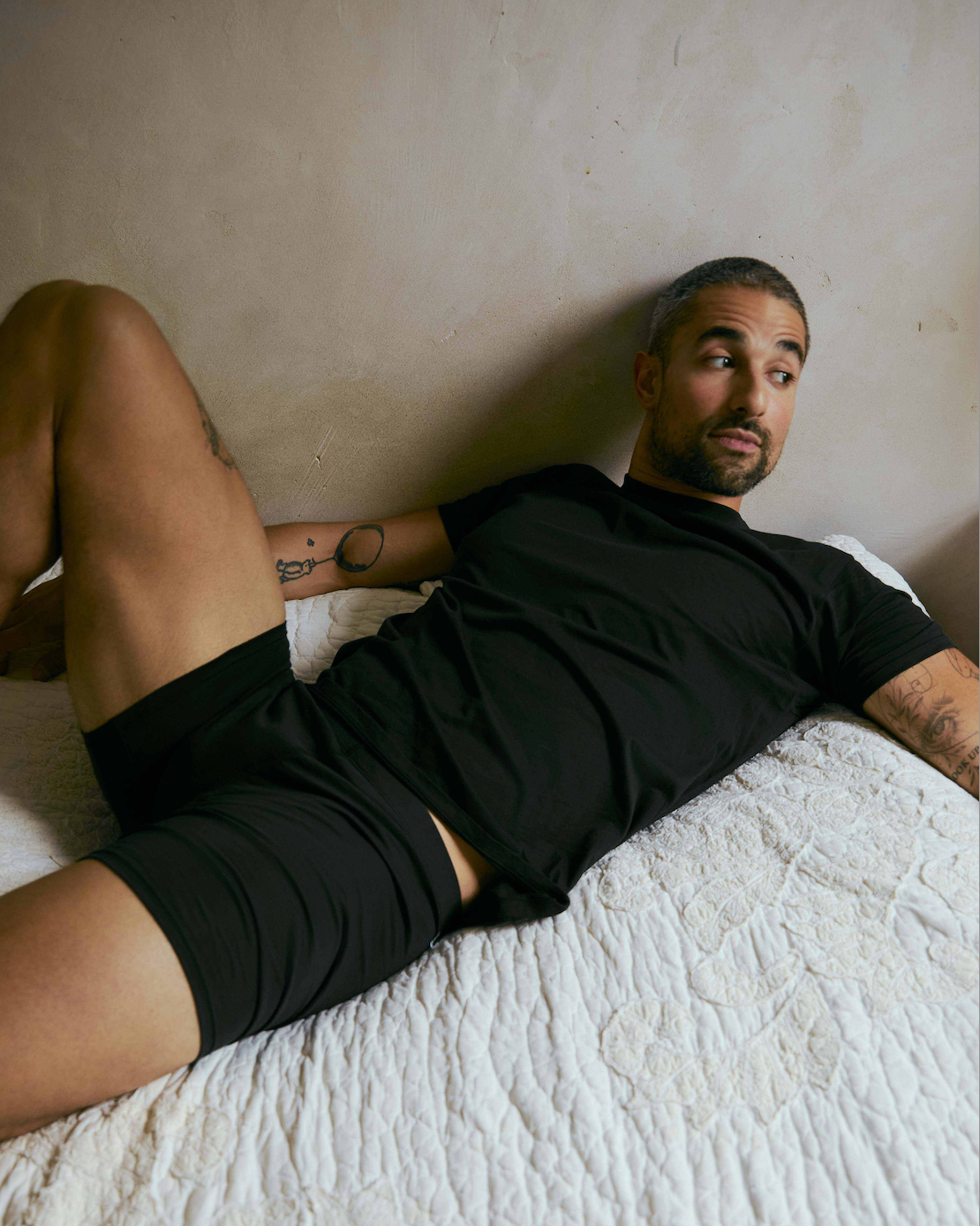 a man wearing the oddobody men's t-shirt  in midnight and oddobody boxer briefs in midnight, reclining on a white textured bed against a neutral beige wall.